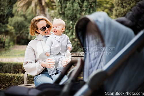 Image of Mother sitting on bench in urban park, laughing cheerfully, holding her smiling infant baby boy child in her lap having baby stroller parked by their site.