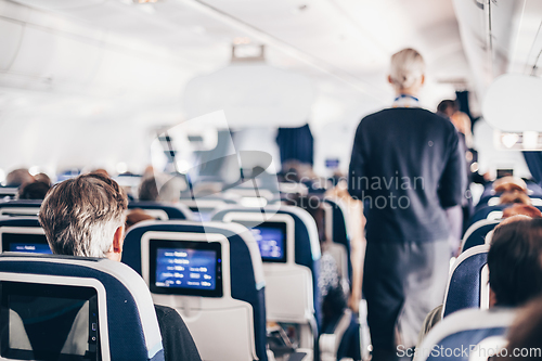 Image of Interior of airplane with passengers on seats and stewardess in uniform walking the aisle, serving people. Commercial economy flight service concept.