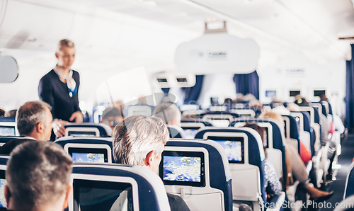 Image of Interior of airplane with passengers on seats and stewardess in uniform walking the aisle, serving people. Commercial economy flight service concept.