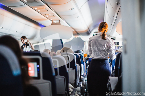 Image of Interior of airplane with passengers on seats and stewardess in uniform walking the aisle, serving people. Commercial economy flight service concept.