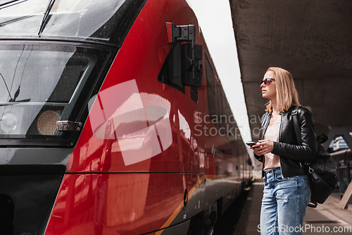 Image of Young blond woman in jeans, shirt and leather jacket wearing bag and sunglass, embarking red modern speed train on train station platform. Travel and transportation.