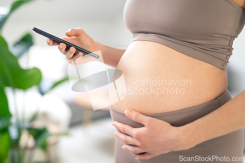 Image of Close-up photo of pregnant female belly. Woman holding and using mobile smart phone application at home interiors. Pregnancy, technology, online shopping, preparation and expectation concept.