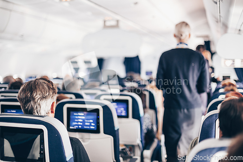 Image of Interior of airplane with passengers on seats and stewardess in uniform walking the aisle, serving people. Commercial economy flight service concept.