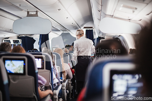 Image of Interior of airplane with passengers on seats and stewardess in uniform walking the aisle, serving people. Commercial economy flight service concept.