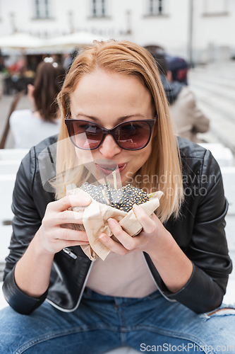 Image of Pretty young blonde funny woman eating hamburger outdoor on the street.