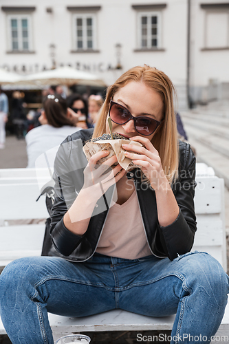 Image of Pretty young blonde funny woman eating hamburger outdoor on the street.