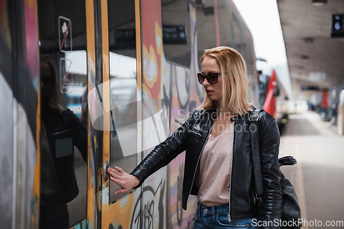 Image of Young blond woman in jeans, shirt and leather jacket wearing bag and sunglass, presses door button of modern speed train to embark on train station platform. Travel and transportation.