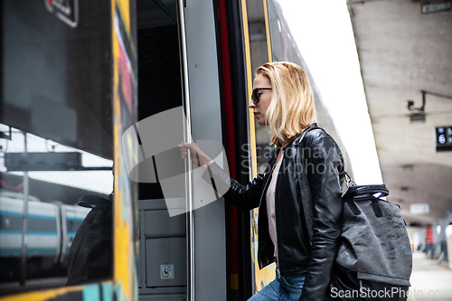 Image of Young blond woman in jeans, shirt and leather jacket wearing bag and sunglass, presses door button of modern speed train to embark on train station platform. Travel and transportation.