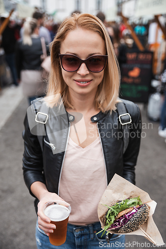 Image of Beautiful young woman holding delicious organic salmon vegetarian burger and homebrewed IPA beer on open air beer an burger urban street food festival in Ljubljana, Slovenia.