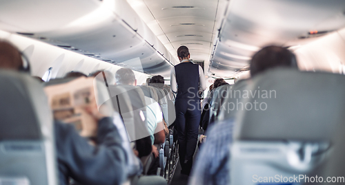 Image of Interior of airplane with passengers on seats and stewardess in uniform walking the aisle, serving people. Commercial economy flight service concept.