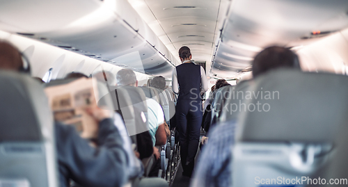 Image of Interior of airplane with passengers on seats and stewardess in uniform walking the aisle, serving people. Commercial economy flight service concept.