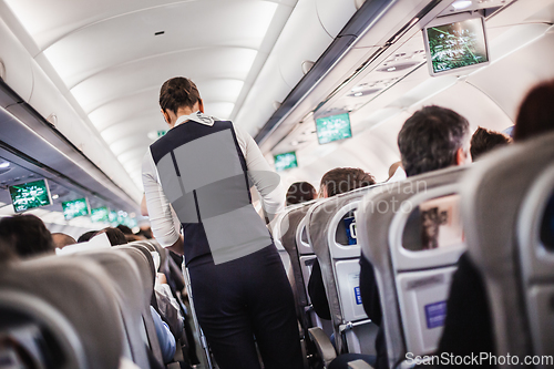 Image of Interior of airplane with passengers on seats and stewardess in uniform walking the aisle, serving people. Commercial economy flight service concept.