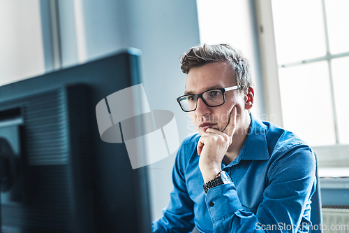 Image of Handsome Young Businessman Wearing Eyeglasses Sitting at his Table Inside the Office, Looking at the Report on his Computer Screen.