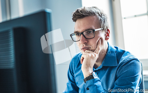 Image of Handsome Young Businessman Wearing Eyeglasses Sitting at his Table Inside the Office, Looking at the Report on his Computer Screen.
