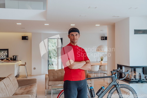 Image of Photo of a determined triathlete standing in a modern large living room, behind him is his training bike