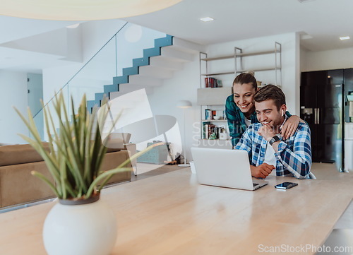 Image of A young married couple is talking to parents, family and friends on a video call via a laptop while sitting in the living room of their modern house