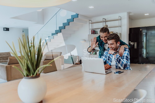 Image of A young married couple is talking to parents, family and friends on a video call via a laptop while sitting in the living room of their modern house