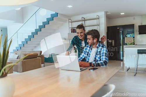 Image of A young married couple is talking to parents, family and friends on a video call via a laptop while sitting in the living room of their modern house