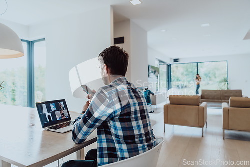 Image of The man sitting at a table in a modern living room, using a smartphone and laptop for business video chat, conversation with friends and entertainment