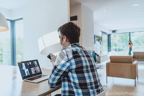 Image of The man sitting at a table in a modern living room, using a smartphone and laptop for business video chat, conversation with friends and entertainment