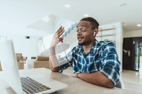 Image of African American man in glasses sitting at a table in a modern living room, using a laptop for business video chat, conversation with friends and entertainment