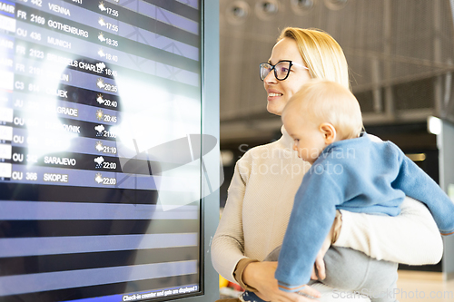 Image of Mother traveling with child, holding his infant baby boy at airport terminal, checking flight schedule, waiting to board a plane. Travel with kids concept.