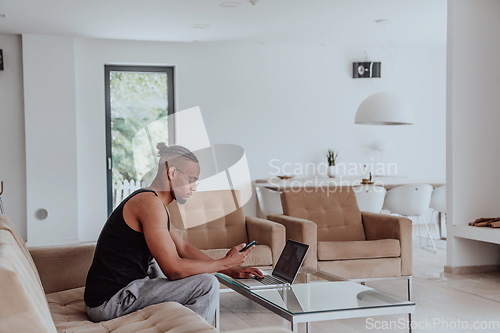 Image of African American sport man in glasses sitting at a table in a modern living room, using a laptop for business video chat, conversation with friends and entertainment