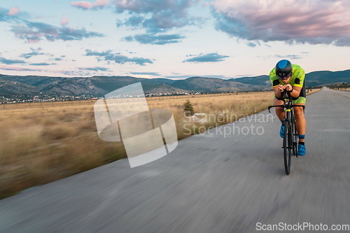 Image of Triathlete riding his bicycle during sunset, preparing for a marathon. The warm colors of the sky provide a beautiful backdrop for his determined and focused effort.