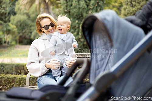 Image of Mother sitting on bench in urban park, laughing cheerfully, holding her smiling infant baby boy child in her lap having baby stroller parked by their site.