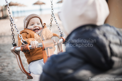 Image of Mother pushing her cheerful infant baby boy child on a swing on sandy beach playground outdoors on nice sunny cold winter day in Malaga, Spain.