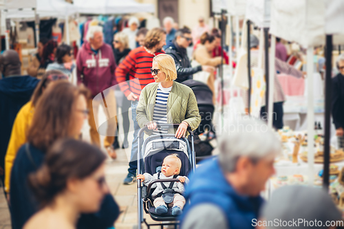 Image of Mother waling and pushing his infant baby boy child in stroller in crowd of unrecognizable people wisiting sunday flea market in Malaga, Spain.