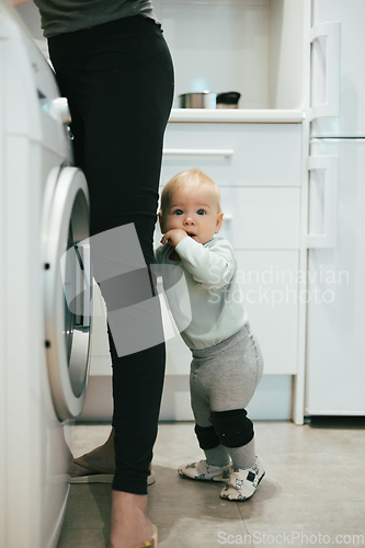 Image of Little infant baby boy child hiding between mothers legs demanding her attention while she is multitasking, trying to do some household chores in kitchen at home. Mother on maternity leave.