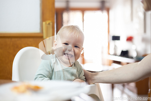 Image of Adorable cheerful happy infant baby boy child smiling while sitting in high chair at the dining table in kitchen at home beeing spoon fed by his mother