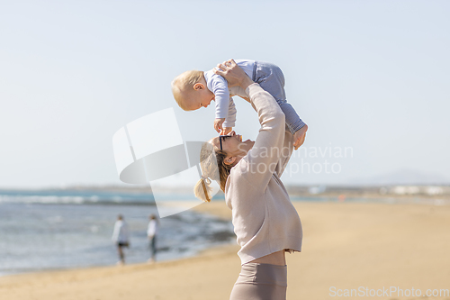 Image of Mother enjoying summer vacations holding, playing and lifting his infant baby boy son high in the air on sandy beach on Lanzarote island, Spain. Family travel and vacations concept.