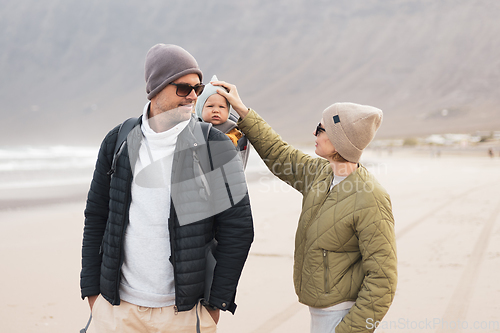 Image of Young father carrying his infant baby boy son in backpack on windy sandy beach. Family travel and winter vacation concept.