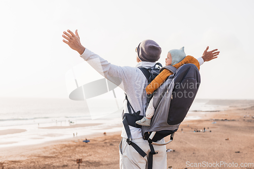 Image of Young father rising hands to the sky while enjoying pure nature carrying his infant baby boy son in backpack on windy sandy beach. Family travel concept.