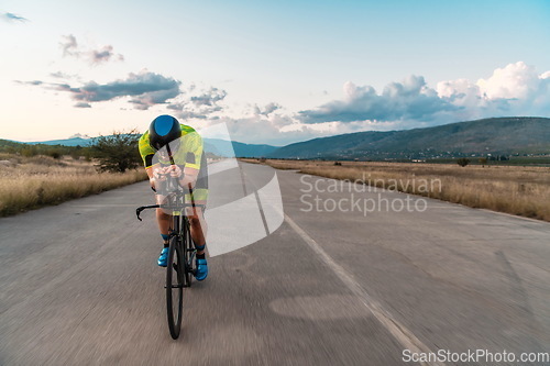 Image of Triathlete riding his bicycle during sunset, preparing for a marathon. The warm colors of the sky provide a beautiful backdrop for his determined and focused effort.