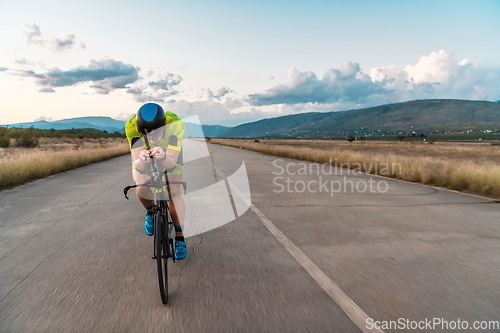 Image of Triathlete riding his bicycle during sunset, preparing for a marathon. The warm colors of the sky provide a beautiful backdrop for his determined and focused effort.