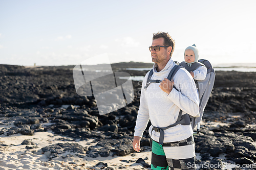 Image of Young father carrying his infant baby boy son in backpack on black rock volcanic beach on Lanzarote island, Spain. Family travel and winter vacation concept.