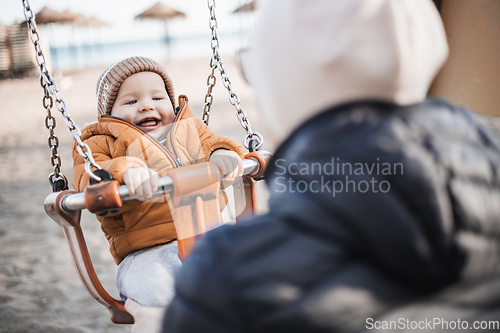 Image of Mother pushing her cheerful infant baby boy child on a swing on sandy beach playground outdoors on nice sunny cold winter day in Malaga, Spain.