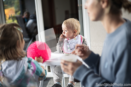 Image of Mother spoon feeding her baby boy child in baby high chair with fruit puree at dinning table at home. Baby solid food introduction concept.