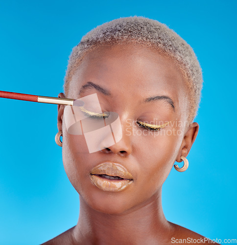 Image of Face, makeup and eyeshadow with a black woman on a blue background in studio to apply a beauty product. Cosmetics, gold color on a brush and a young female model eyes closed for style or glamour