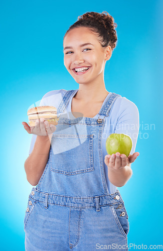 Image of Portrait, smile and woman with burger, apple and choice in studio isolated on a blue background. Face, fruit and person with sandwich, fast food or decision for healthy diet, nutrition or wellness.