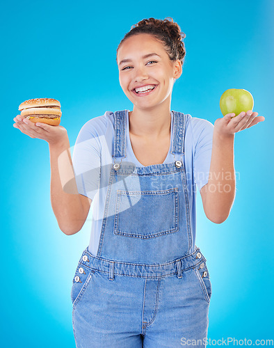 Image of Happy, portrait and woman with burger, apple and choice in studio isolated on a blue background. Smile, fruit and person with sandwich, fast food or comparison for healthy diet, nutrition or wellness