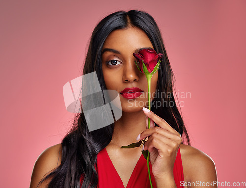 Image of Face, beauty and portrait of a woman with a rose on a studio background for valentines day. Makeup, model and serious young Indian girl with a flower in hand for romance or love on pink backdrop