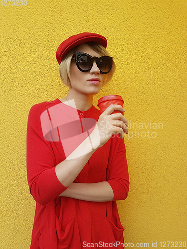 Image of Female model in long red sweater and red hat with red paper cup standing on yellow background. Outdoor photo of beautiful caucasian woman in sweater drinks coffee in front of cafe