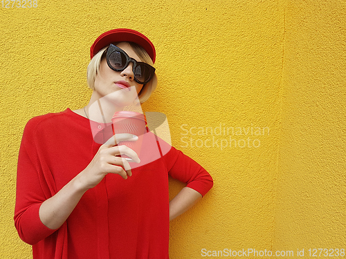 Image of Female model in long red sweater and red hat with red paper cup standing on yellow background. Outdoor photo of beautiful caucasian woman in sweater drinks coffee in front of cafe