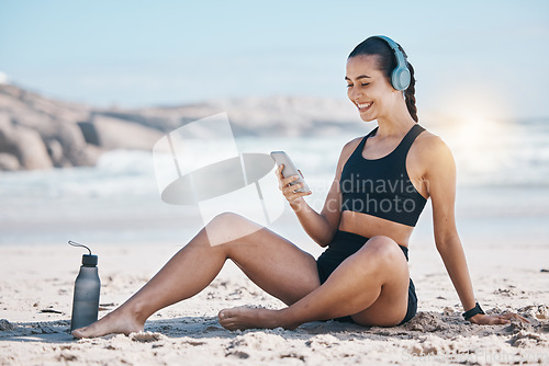 Image of Fitness, break and happy woman relax at beach with phone, music and headphones after training. Exercise, resting and lady health influencer posting to social media, blog or podcast after ocean run