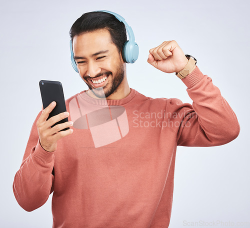 Image of Man, headphones and celebrate success with smartphone, music and white background in studio. Asian male model, cellphone and fist for happy dance, excited audio deal and hearing winning news on radio