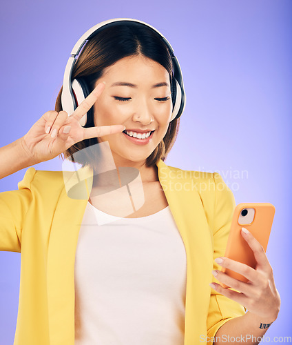 Image of Peace sign, selfie and happy Asian woman in studio smile for social media, online post or mobile app. Headphones, music and female person with emoji on smartphone listen to audio on purple background